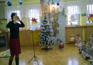Świątecznie udekorowana sala, śpiewaczka śpiewa kolędę. Dzieci podziwiają śpiew.
